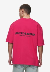 Túlméretezett rózsaszín pamut póló, fekete grafikával ellátott "PEGADOR" felirattal a hátsó részén, rövid ujjakkal és laza szabással.