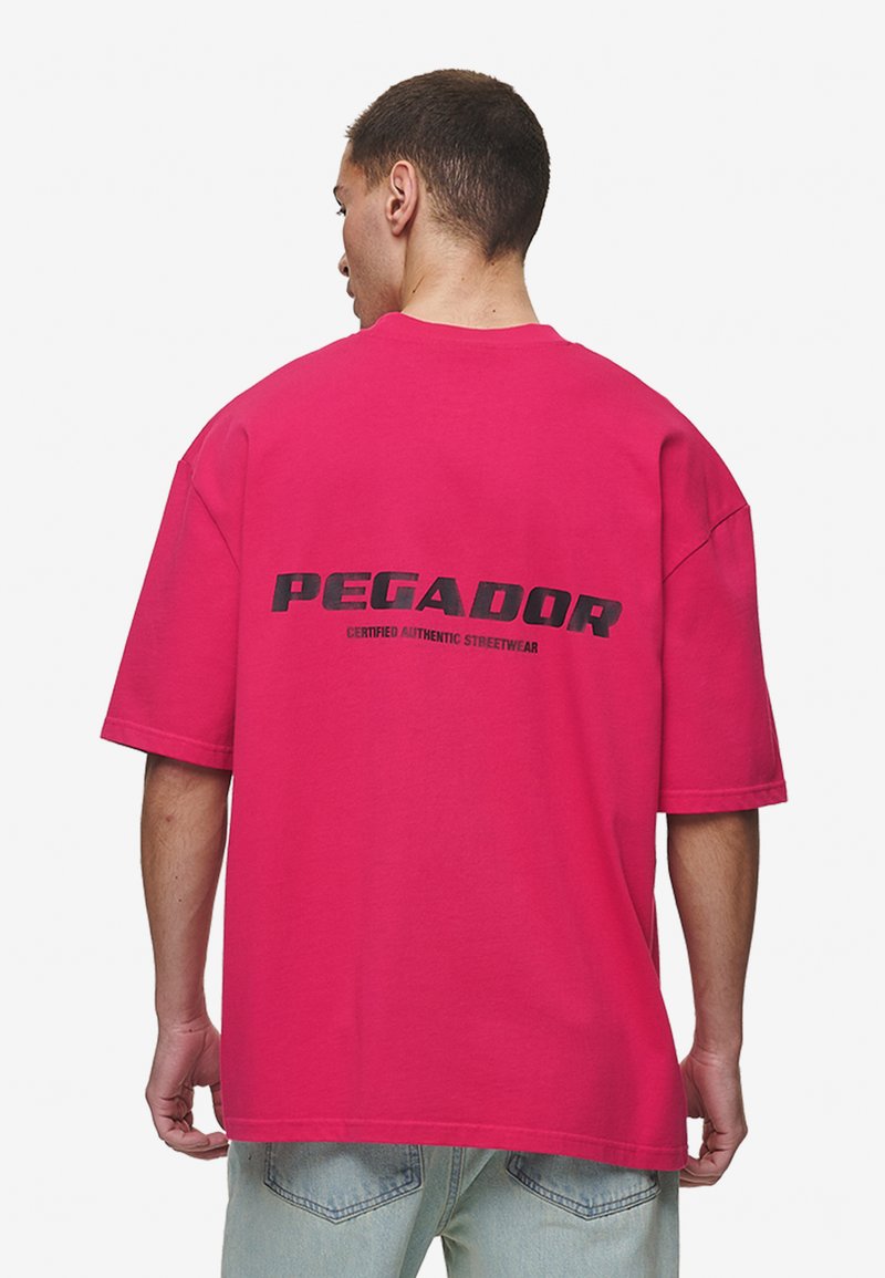 Túlméretezett rózsaszín pamut póló, fekete grafikával ellátott "PEGADOR" felirattal a hátsó részén, rövid ujjakkal és laza szabással.