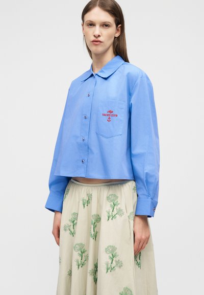 Νεαρή γυναίκα που φοράει κοντή μπλε πουκάμισο με κουμπιά και κόκκινη κεντημένη επιγραφή «Yacht Club» και κρεμ φούστα με πράσινα λουλουδένια σχέδια.
