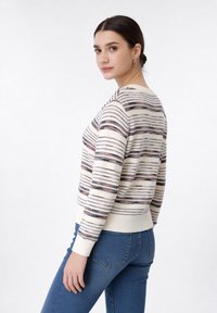 Jeune femme aux cheveux foncés portant un pull crème à rayures et un jean bleu, debout de profil et regardant vers la caméra sur un fond blanc.