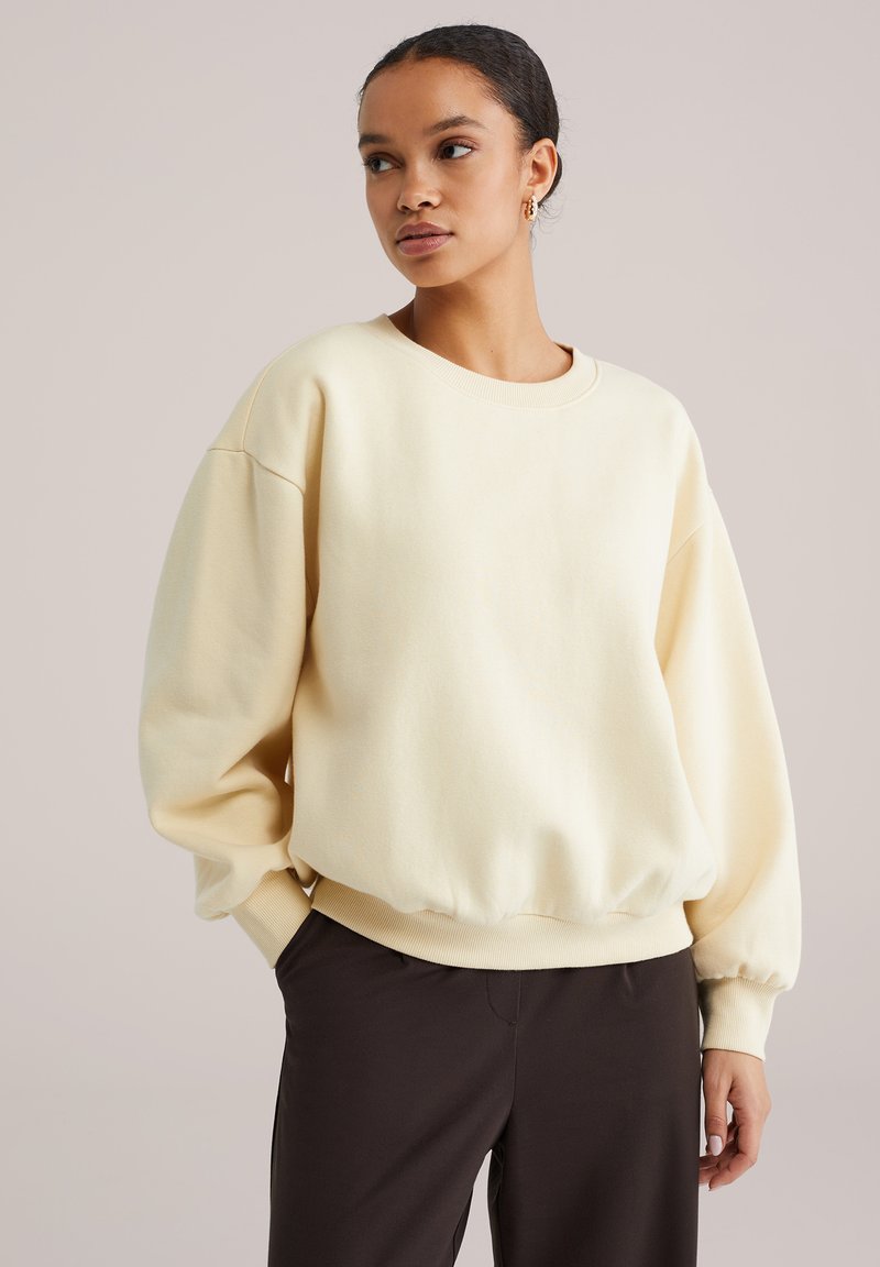 Kreemika värvi dressipluus ümmarguse kaelusega, langevate õlgade ja soonikkoes varrukate äärega. Pehme tekstuuriga ja lõdva istuvusega. Kandmiseks tumedate pükstega.