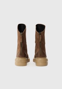 Bottes courtes en cuir marron avec une fermeture éclair à l'arrière, un bout arrondi et une semelle en caoutchouc texturée de couleur beige. L'intérieur est doublé de noir.