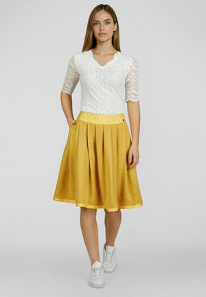 Vrouw die loopt, gekleed in een witte kanten top met halve mouwen, een mosterdgele geplooide rok met een bloemenriem en witte sneakers, tegen een grijze achtergrond.