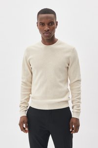 Beige gebreide trui met een ronde halslijn, lange mouwen, geribde manchetten en zoom, gecombineerd met zwarte broek, tegen een lichte achtergrond.