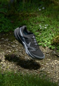Chaussure de sport noire avec une tige en maille texturée, accents gris et semelle rembourrée, flottant au-dessus d'un sol rocheux et de zones herbeuses.