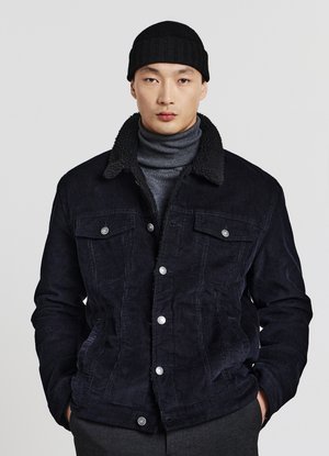 Uomo in giacca di corda nera con colletto in pile, dolcevita grigia, berretto nero e pantaloni scuri, in piedi contro uno sfondo luminoso e semplice.