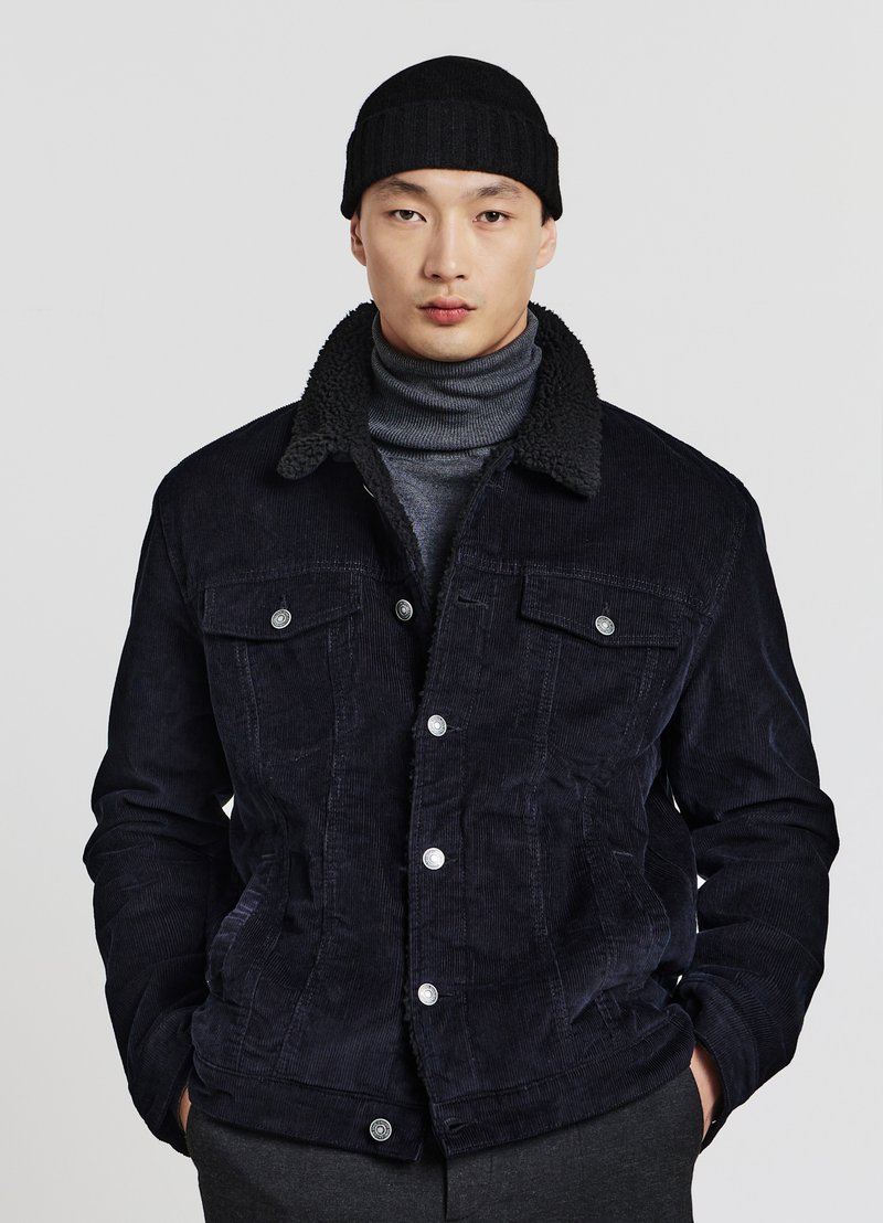 Mann trägt eine schwarze Cordjacke mit Fleecekragen, einen grauen Rollkragenpullover, eine schwarze Mütze und dunkle Hosen, steht vor einem einfachen hellen Hintergrund.