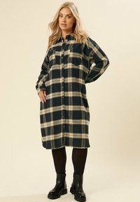 Czarna i beżowa koszulowa sukienka w kratę z długimi rękawami, z przodu zapinana na guziki, z kołnierzykiem i jedną kieszenią na piersi. Stylizowana z czarnymi botkami za kostkę.