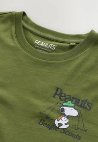 Vihreä puuvillapaita, jossa on grafiikka Snoopy-hahmosta hatussa tekstillä "Peanuts Beagle Scouts" ja leirintäelementtejä. Pyöreä pääntie.