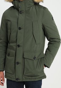 Parka isolante verte avec capuche bordée de fourrure, fermeture à boutons, quatre poches et coupe décontractée, fabriquée dans un tissu durable.