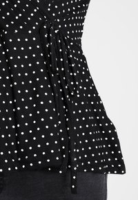 Svart tygtop med vita prickar, med en knytdetalj i midjan och en utsvängd silhuett. Slät textur, avslappnad design.