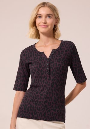 Femme blonde portant un haut violet foncé à motif léopard avec des boutons et des manches aux coudes, souriant légèrement sur un fond neutre.