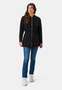 Schwarze Zip-Hoodie mit Kordelzug in der Taille, langen Ärmeln und Kapuze. Trage sie über blauen Jeans und weißen Sneakers. Einfaches Design, keine Muster.