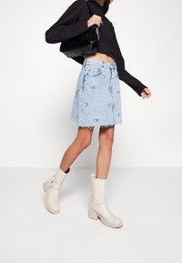 Denim skirt in light blue with embroidered logo patches, frayed hem, paired with a black crop top and cream-colored chunky heeled boots.