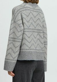 Femme portant un pull en tricot gris avec un motif en zigzag et des poignets côtelés, associé à un pantalon gris foncé, vue de dos.