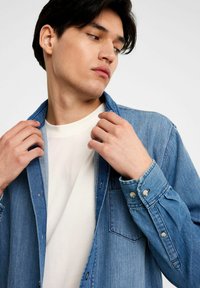 Jasnoniebieska koszula denimowa z guzikami, z przednią kieszenią, zawiniętymi rękawami i miękkim białym t-shirtem pod spodem. Gładka faktura, casualowy styl.