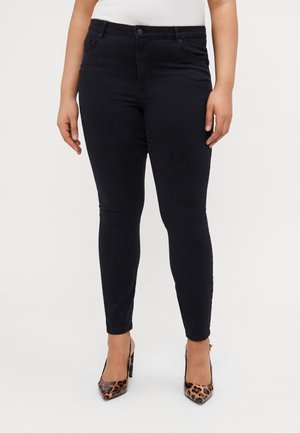 Vrouw draagt zwarte skinny jeans en hoge hakken met luipaardprint, staand tegen een effen lichte achtergrond.