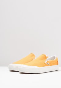Orangefarbene Canvas-Slipper mit weißer Gummisohle und strukturierter Naht. Verfügt über ein kleines Logetikett an der Seite.