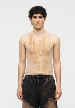 Νέος άνδρας με σγουρά μαλλιά που φοράει αμάνικο μπεζ διχτυωτό τοπ με υφή κάθετες ρίγες και άνετο μαύρο παντελόνι με σχέδιο.