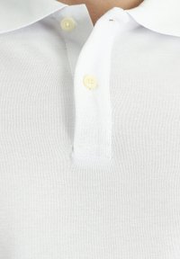 Polo bianca con una texture morbida, dotata di un colletto classico e due bottoni gialli all'apertura del collo. Nessun motivo o dettaglio visibile.