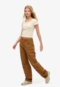 Top de punto de color beige, de manga corta y con cierre de botones, combinado con pantalones cargo de algodón marrón, holgados, que presentan bolsillos y detalles de costura.