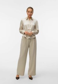 Beige satin bluse med knap-front, lange ærmer og buet kant, parret med beige højtaljede bukser med brede ben og brune spidse sko.