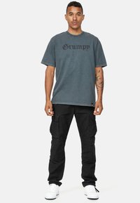 Camiseta gris de gran tamaño con estampado en negro de "Grumpy", combinada con pantalones cargo negros y zapatillas blancas. La tela parece suave con costuras visibles.