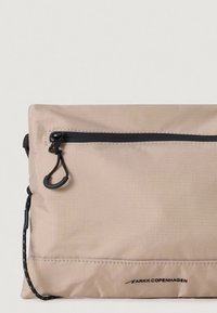 Borsa in tessuto beige con cerniera nera e tracolla regolabile, presenta una superficie testurizzata e un logo del marchio sottile nella parte inferiore.