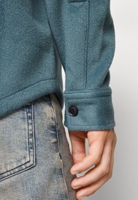 Manche de manteau en laine bleu sarcelle avec détail de bouton noir, portée sur un jean en denim bleu clair délavé, montrant une main détendue à côté de la poche.
