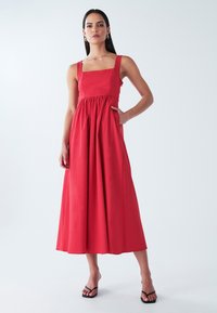Rotes, ärmelloses Maxikleid mit quadratischem Ausschnitt, geraffter Taille und Seitentaschen. Glatte Textur, fließender Stoff und lange Silhouette.