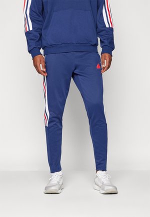 Homme portant un pantalon de sport bleu avec le logo rouge Adidas et des chaussures blanches, debout devant un fond clair uni.