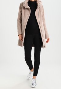 Cappotto marrone chiaro fino al ginocchio con cappuccio, dotato di chiusura a zip e tasche laterali, indossato sopra una maglia nera e leggings, abbinato a sneakers argento.