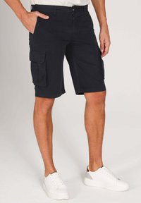 Shorts cargo bleu marine foncé avec deux poches latérales, en tissu mélangé de coton. Dotés d'une fermeture à glissière et d'un bouton. Portés avec des baskets blanches.