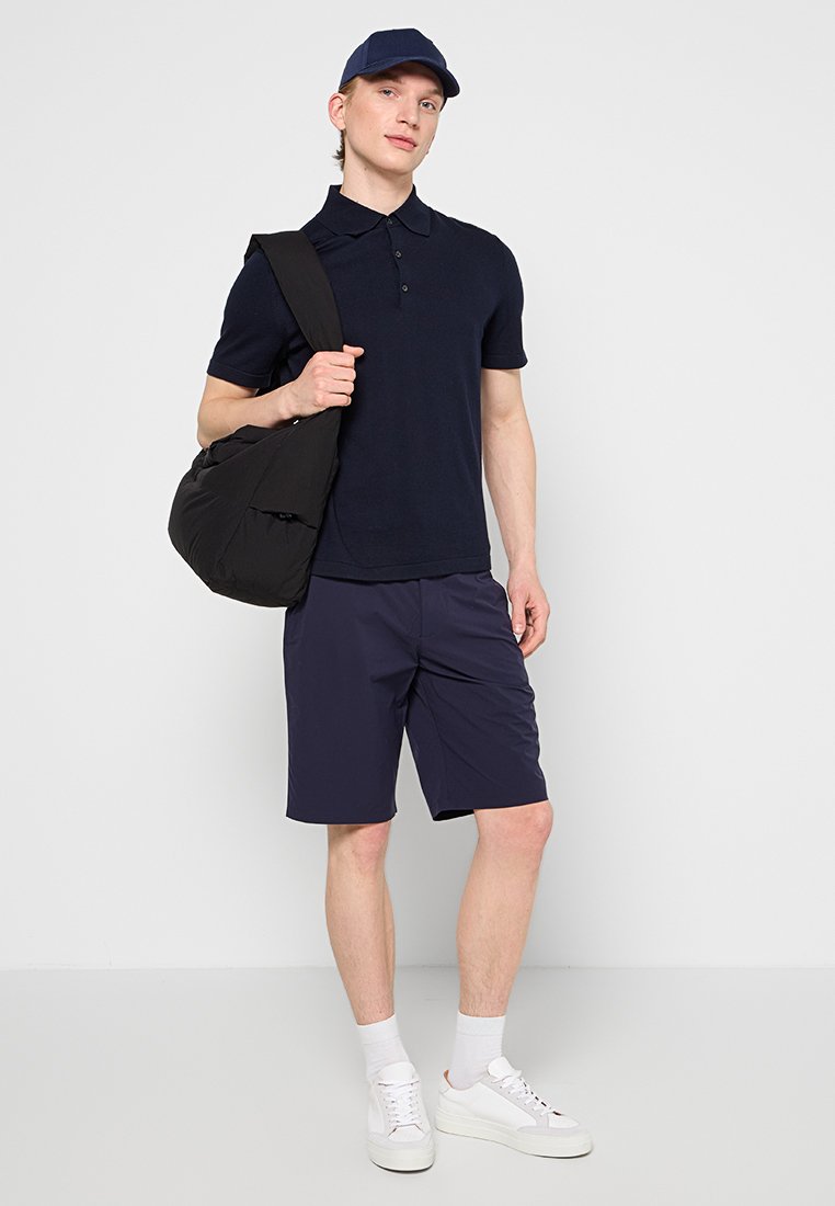 Granatowy polo, ciemne szorty, białe trampki. Wyposażony w czarną torbę na ramię. Model stoi na neutralnym tle.