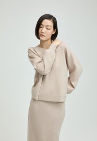 Beige Strickpullover mit langen, leicht gepufften Ärmeln; strukturierter Stoff; lockere Passform; kombiniert mit einem passenden, knallengen Rock. Schlichte Silhouette.