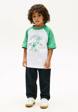 Niño pequeño con cabello rizado que lleva una camiseta deportiva Tommy blanca y verde, vaqueros oscuros y zapatillas blancas, de pie sobre un fondo liso.