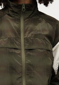 Κοντινό πλάνο ενός ατόμου που φοράει μια ελιάς πράσινη ζακέτα με φερμουάρ και σχέδιο σε μορφή πλέγματος, με μικρό λογότυπο Puma στο στήθος.