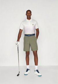 Polo branca com um logotipo amarelo, calções verdes, cinto azul, segurando um taco de golfe, calçando ténis brancos com detalhes azuis.