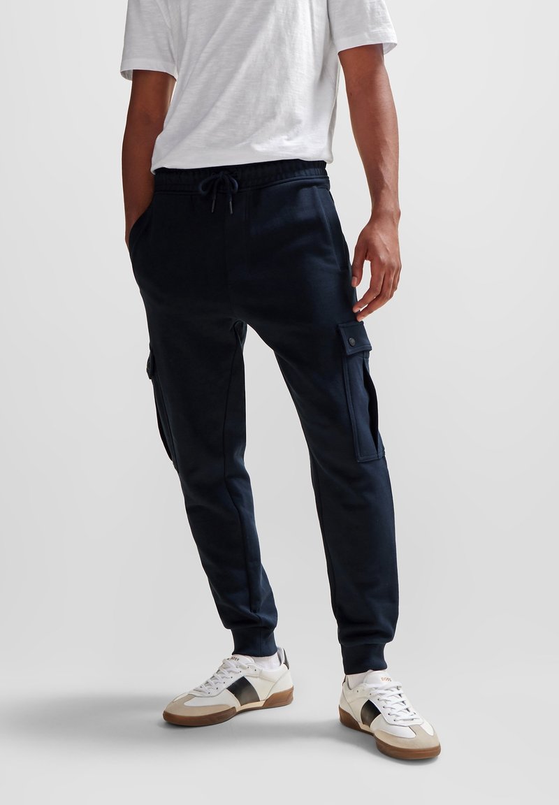 Marinblå sweatsbyxor gjorda av mjukt tyg, med sidofickor i cargo-stil, dragsko i midjan, smalnande ben och ribbade muddmar.