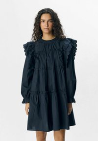 Schwarzes Baumwollkleid mit Rüschenschultern, langen Ärmeln und gestuftem Design. Verfügt über elastische Bündchen und einen gerafften Tailleneinsatz für eine fließende Silhouette.