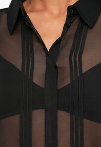 Blusa preta transparente com gola abotoada, apresentando pregas verticais e detalhes opacos na área do busto. Textura lisa, design elegante.