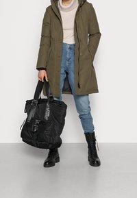 Olivgrön jacka med huva, beige tröja, ljusblå jeans och svarta kängor. Svart tote-väska med texturerad yta och justerbara remmar.