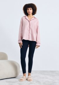 Ljusrosa knapp-up pyjamaströja med röd kant, ihop med marinblå leggings. Modell bär beige fluffiga tofflor och står mot en ljus bakgrund.