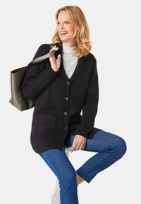 Dunkler Strickcardigan mit Schalkragen, sechs Knöpfen und zwei vorderen Taschen, kombiniert mit blauen Jeans und einem hellen Rollkragenpullover.