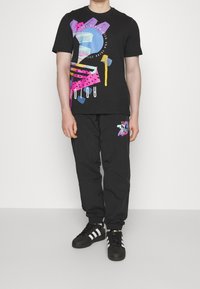 Černé tričko s barevnými abstraktními vzory, doplněné černými kalhotami se stejným motivem. Černé tenisky s bílými pruhy.