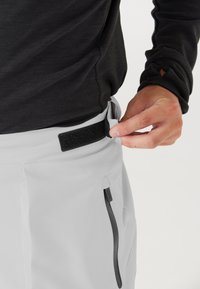 Lichtgrijze sportbroek met een klittenband tailleband, voorzien van een ritszak en zwarte accenten. De stof lijkt glad en rekbaar te zijn.