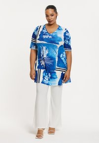 Blaue Blumentunika mit V-Ausschnitt und kurzen Ärmeln, kombiniert mit weißen, ausgestellten Hosen. Das Design zeigt abstrakte Formen und Streifen.