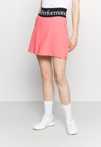 Vaaleanpunainen urheiluhame mustalla vyötärökaistalla, jossa on "Peak Performance" -logo. Hameessa on sivuaukot liikkuvuutta varten, ja se on yhdistetty valkoisiin lenkkikenkiin.