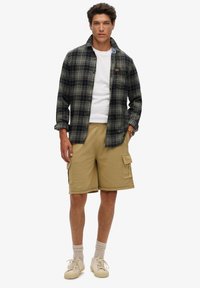 Camisa de franela en cuadros azul marino y verde con un bolsillo en el pecho, combinada con pantalones cortos de cargo color caqui, camiseta blanca y zapatillas color beige.
