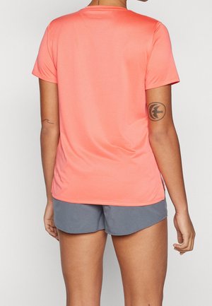 Person iført koralfarvet t-shirt med korte ærmer og grå shorts, vist bagfra, med arm-tatoveringer synlige på overarmene.